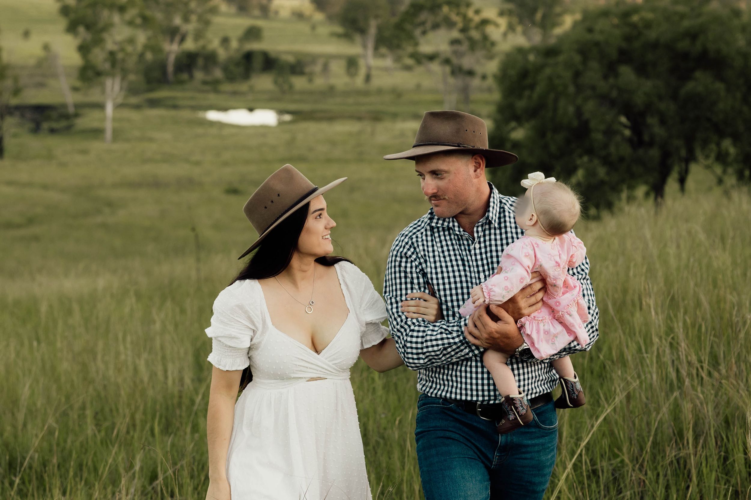 A couple in cowboy hats walk through green fields, man holding baby girl in pink dress, woman in white dress.