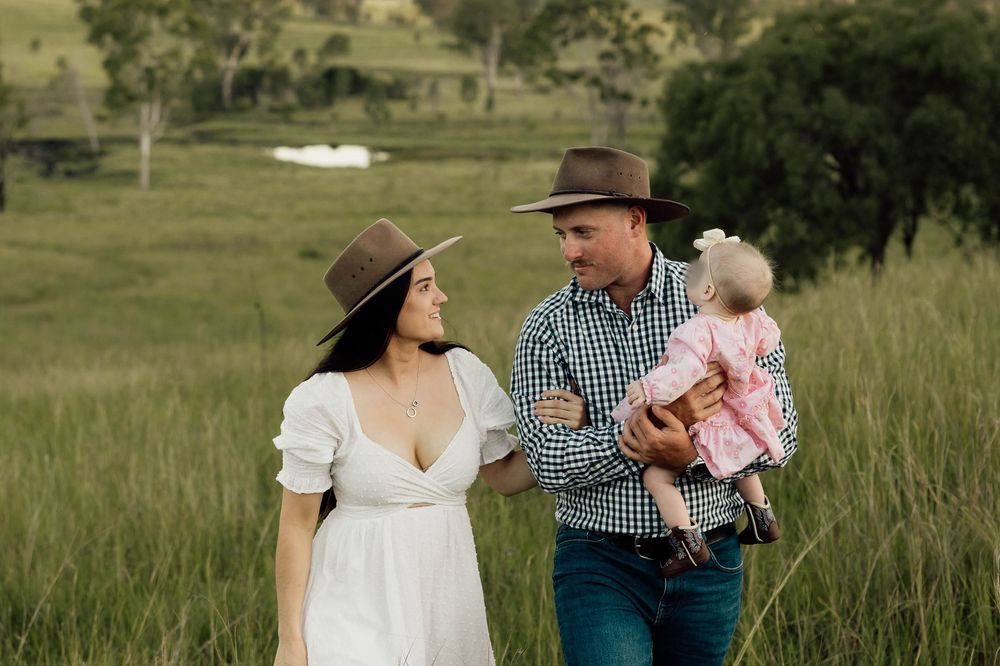 A couple in cowboy hats walk through green fields, man holding baby girl in pink dress, woman in white dress.