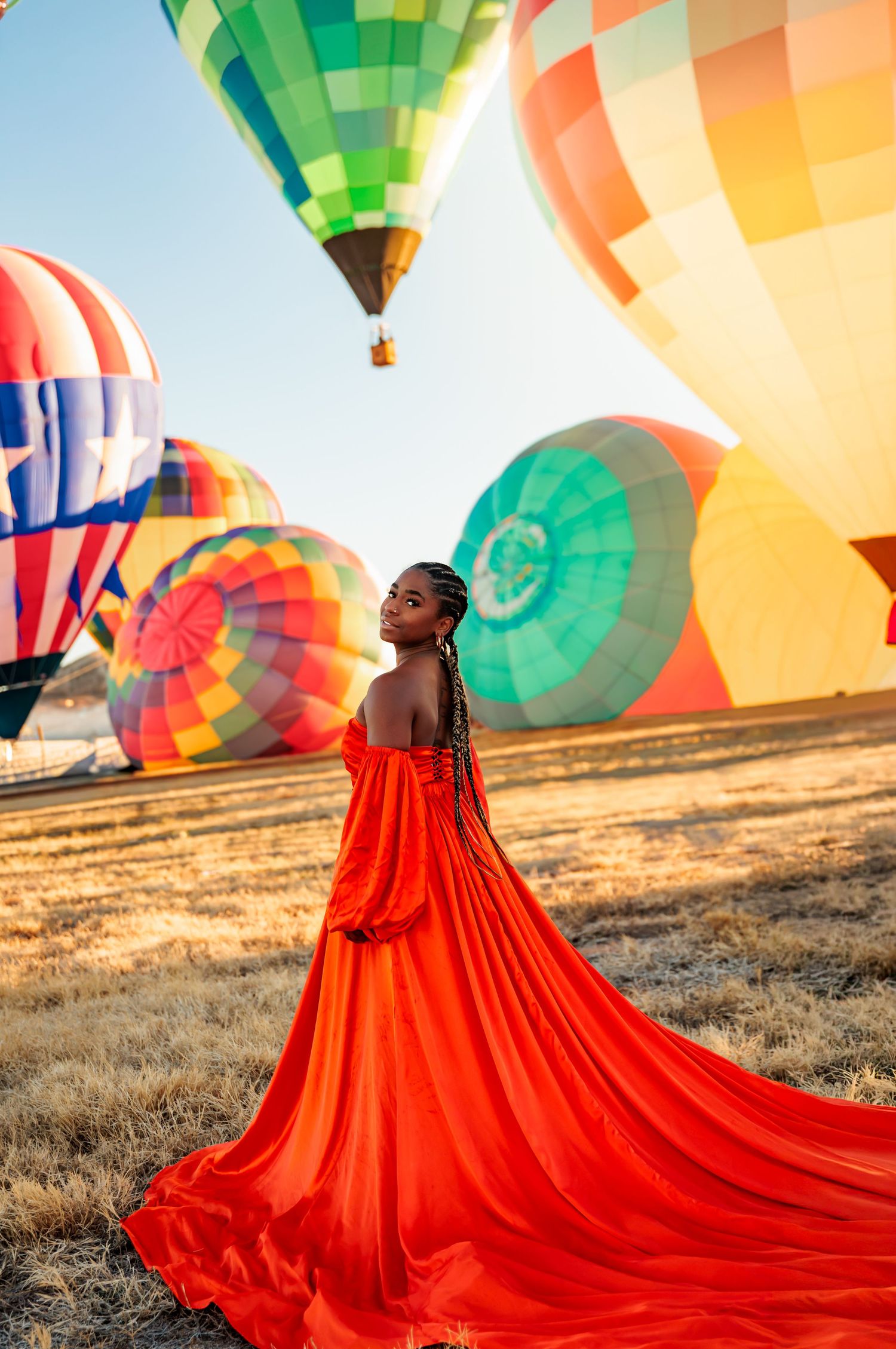 Woman in flowing red gown poses at hot air balloon festival with colorful balloons launching at golden hour.