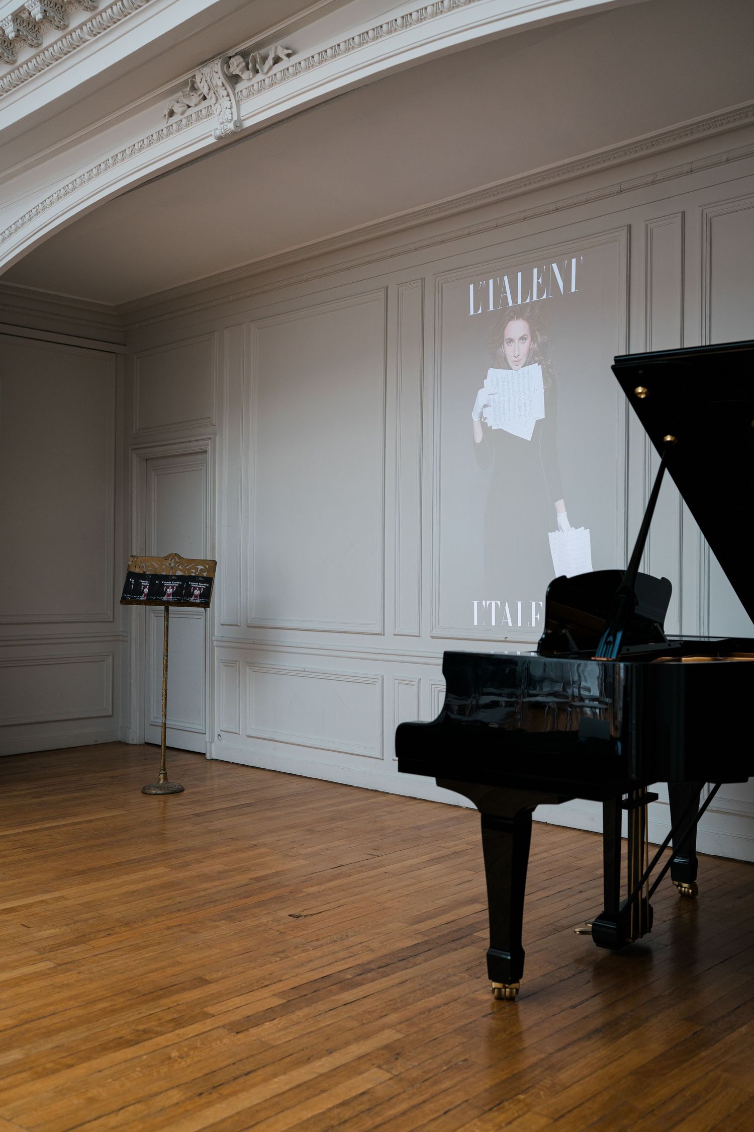 Elegant concert hall with a black grand piano, music stand, and L'Taleni magazine projected on a white paneled wall.