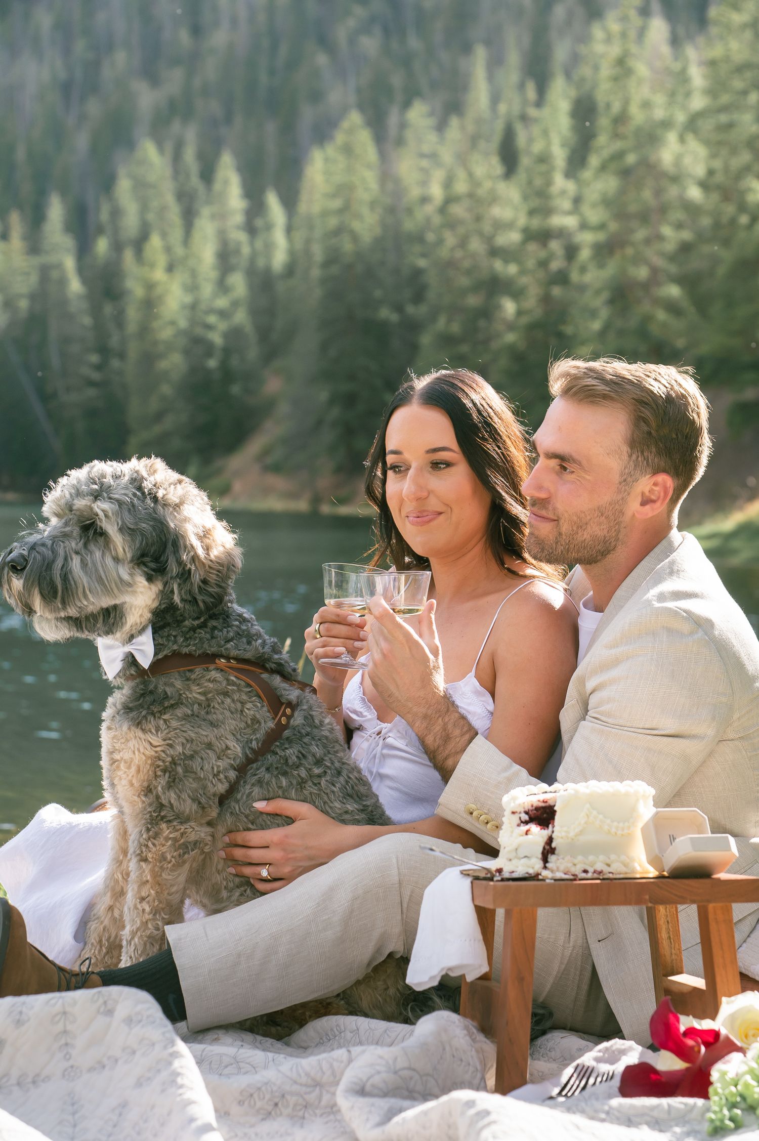 Couple toasting with champagne at a lakeside picnic with their fluffy dog for a Colorado mountain elopement at Officer's Gulch