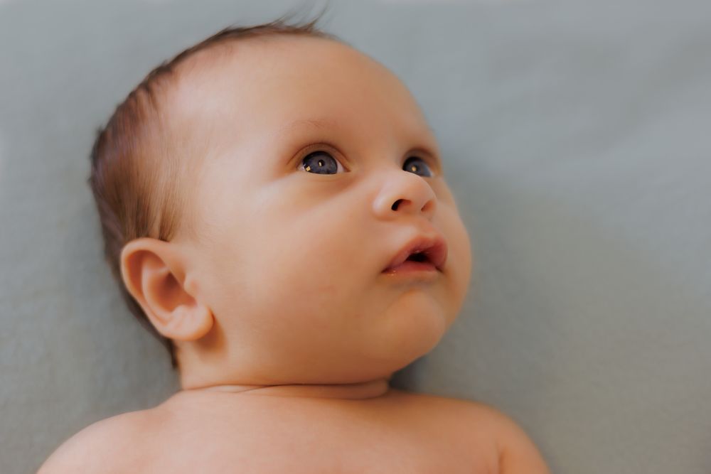 Close-up portrait of an adorable newborn baby lying on a gray blanket, looking up with curious blue eyes.