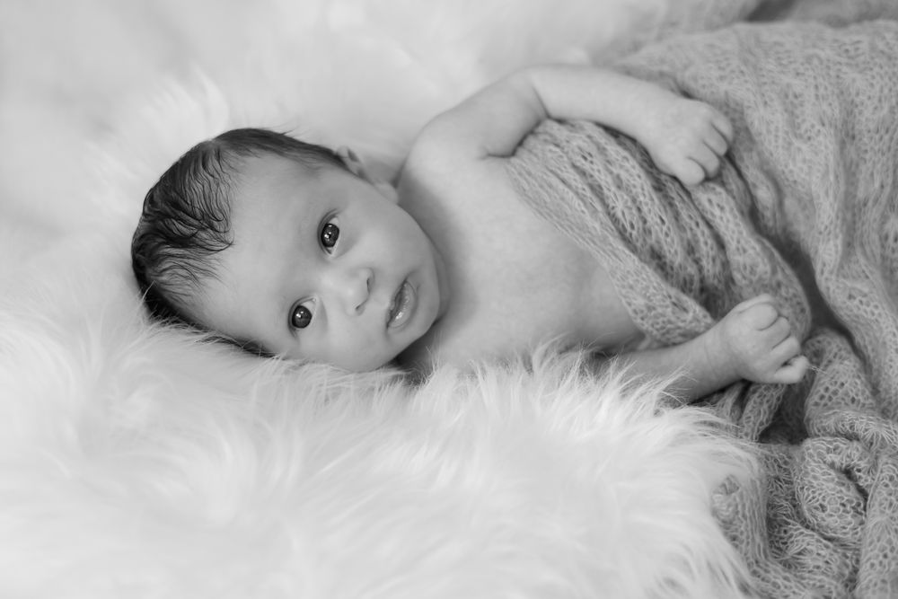 Black and white photo of newborn baby wrapped in knit blanket resting on fluffy white fur, gazing softly.