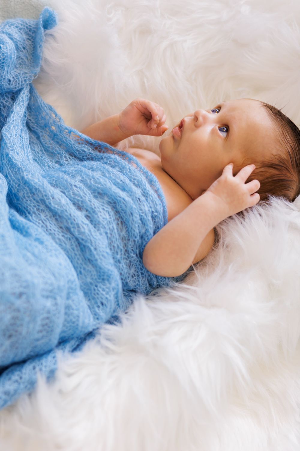 Newborn baby wrapped in a soft blue knit blanket lying on a white fluffy fur rug, looking upward.
