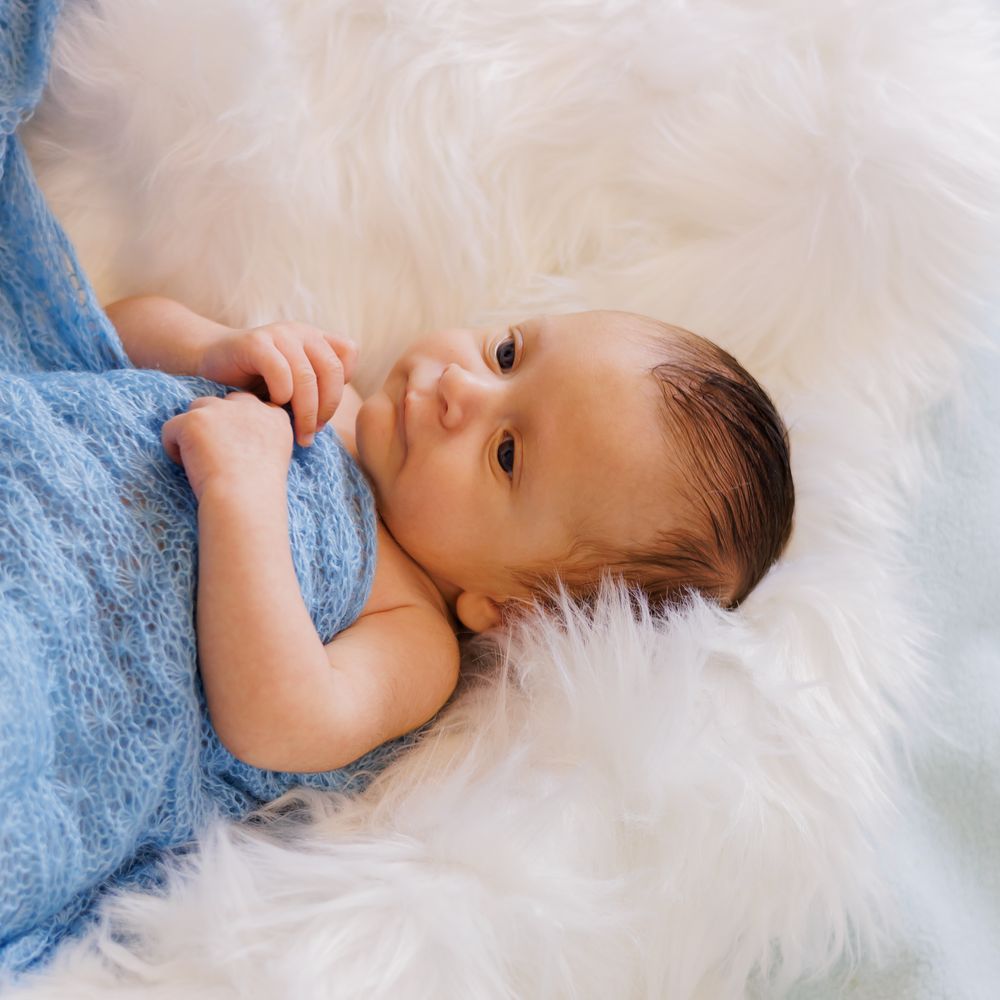 Newborn baby wrapped in blue knit blanket lying on fluffy white fur, gazing upward in a serene portrait.