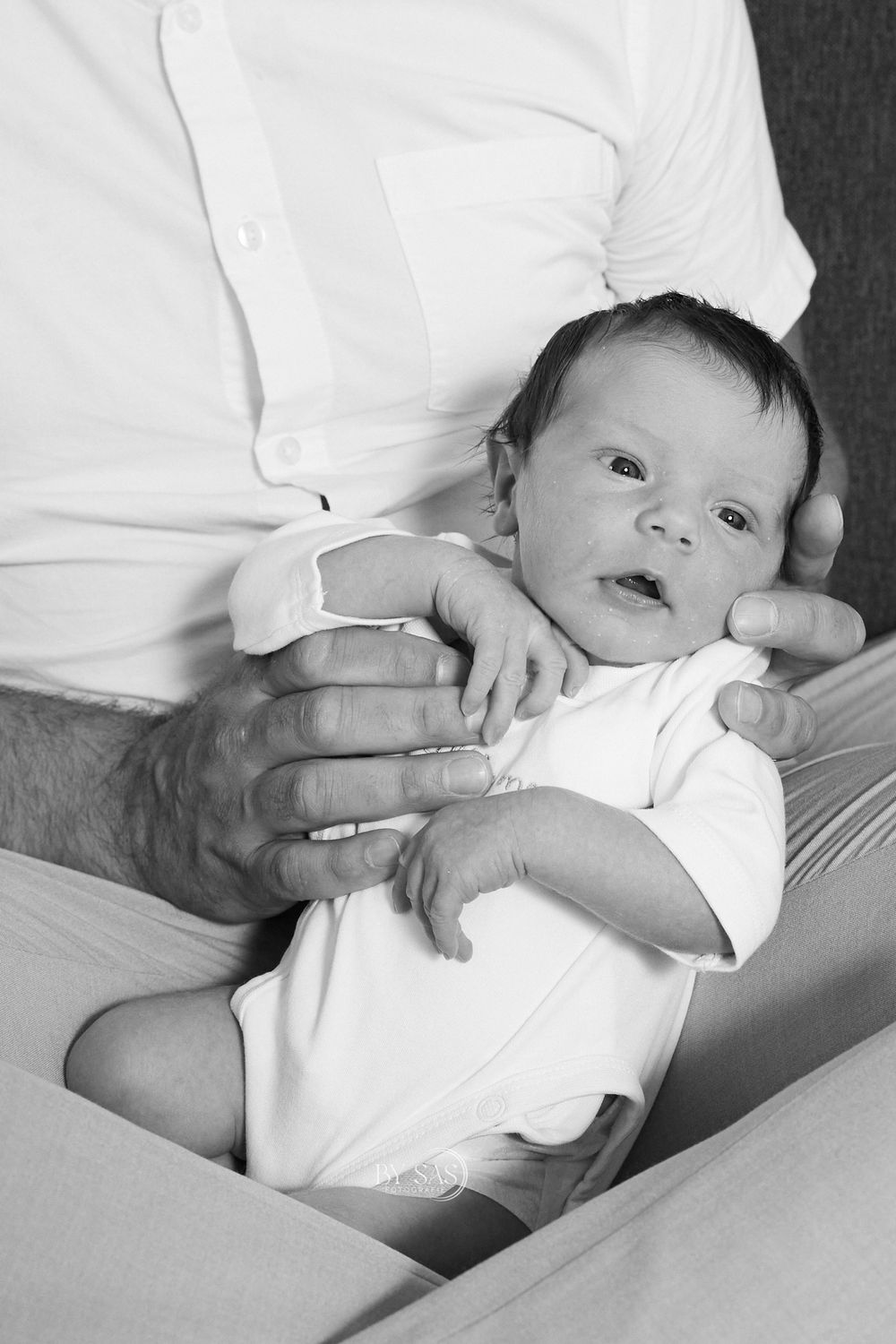 Black and white photo of a newborn baby in white onesie being held by an adult's hands.