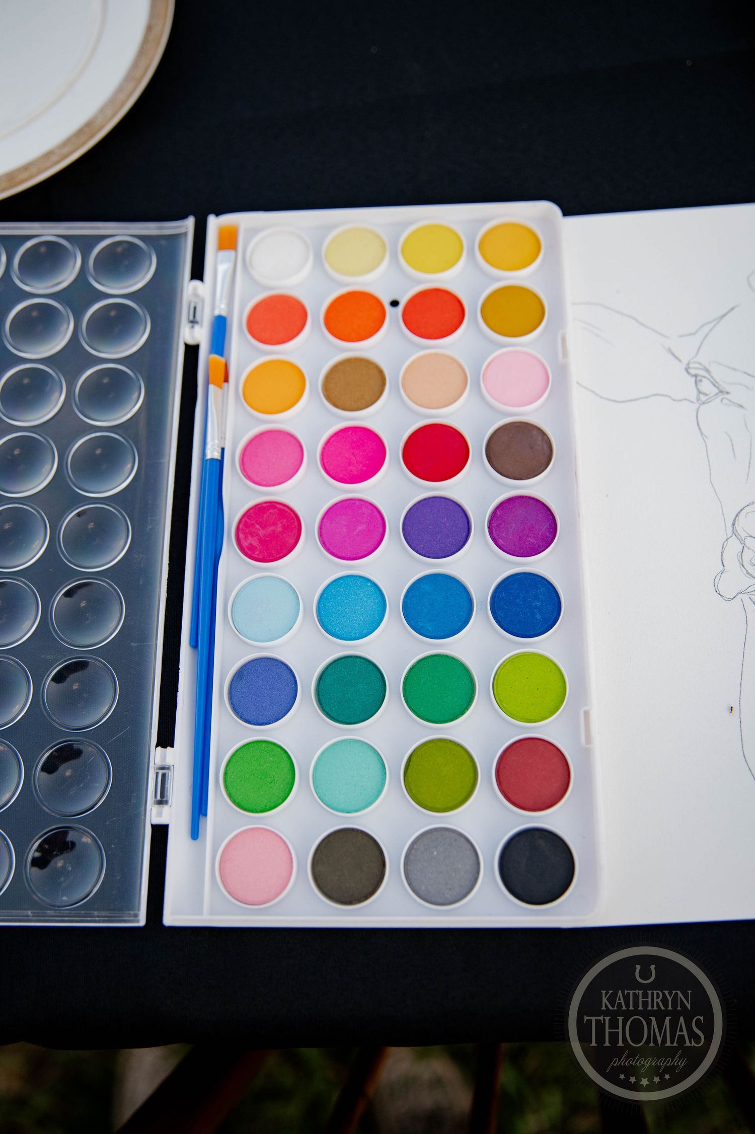 Vibrant watercolor paint palette with 36 colorful pans, two blue brushes, and a sketch pad on a dark table.