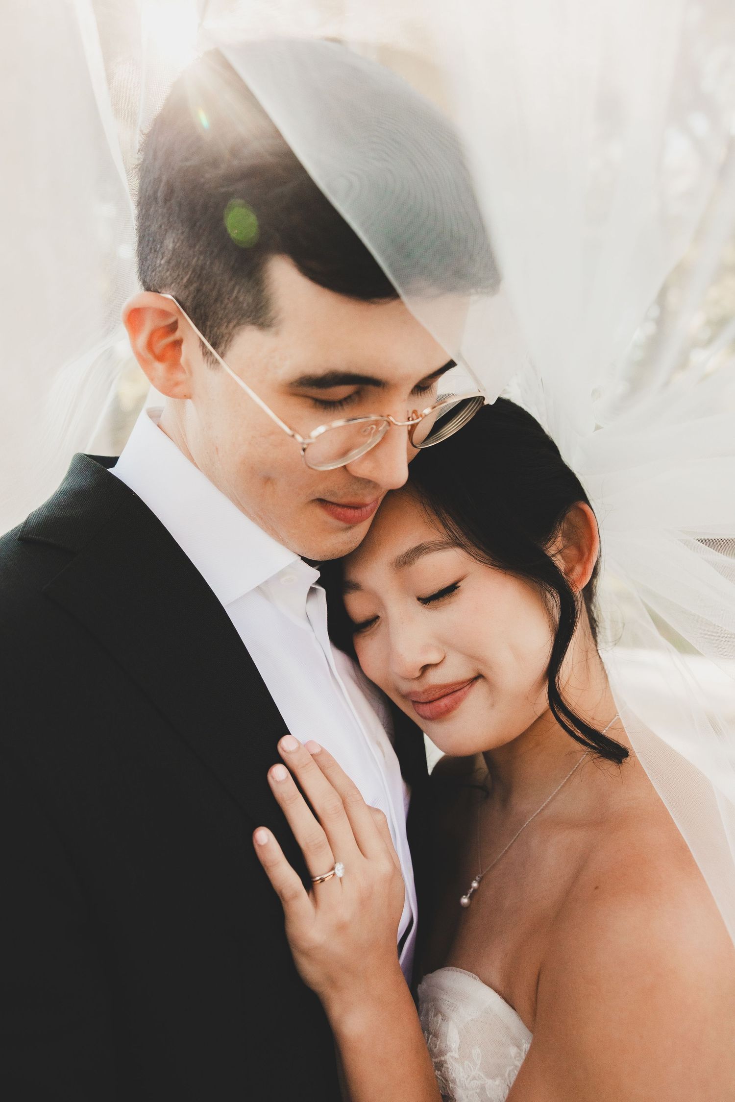Romantic wedding couple embracing under a veil, groom in black suit with glasses, bride in white dress with eyes closed.