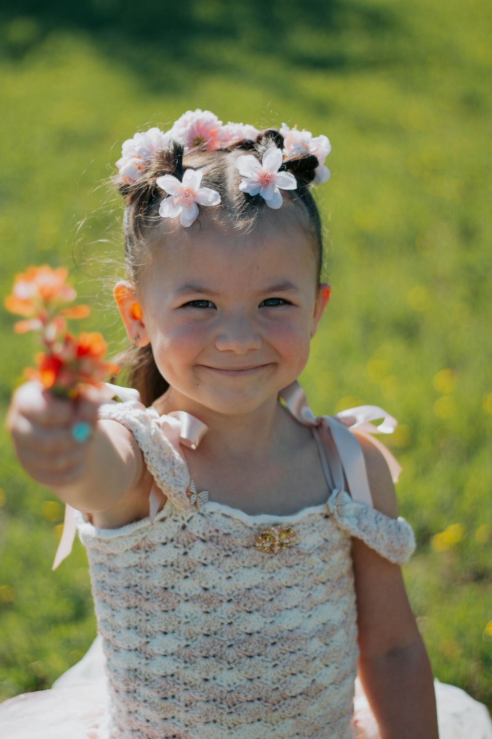 Smiling little girl in crochet dress with floral crown holding out wildflowers in a sunny green meadow.