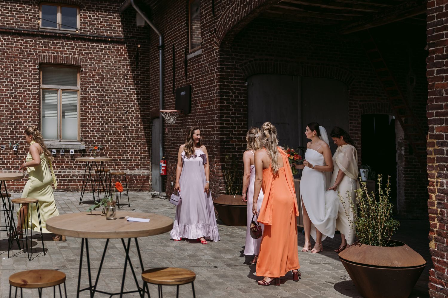 Bridesmaids in colorful dresses gather in a brick courtyard during an outdoor wedding reception.