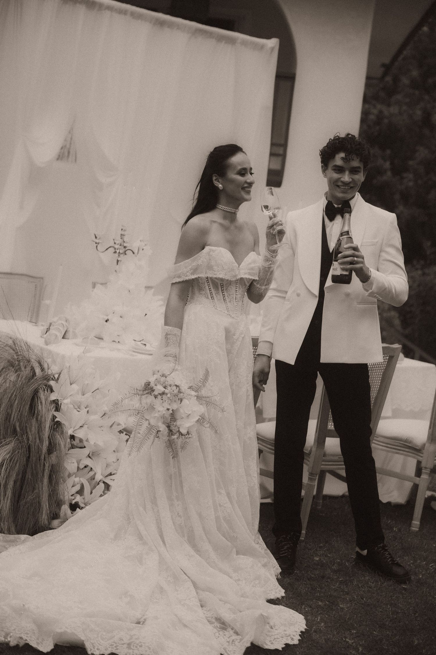 Bride and groom toasting with champagne at outdoor wedding reception, bride in lace gown holding bouquet.