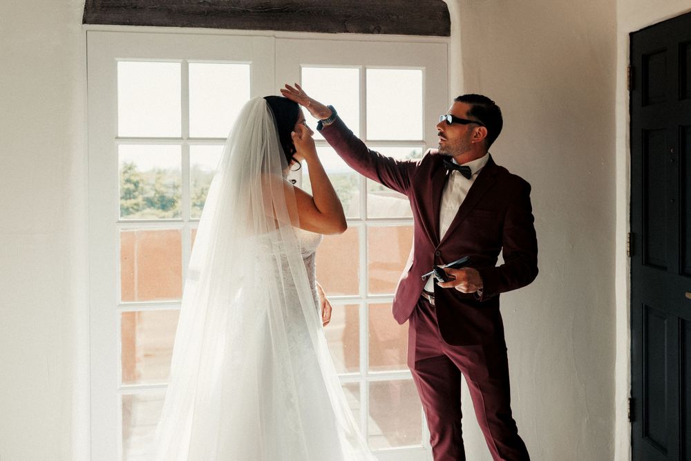 Groom in burgundy suit adjusting bride's veil by window on wedding day.