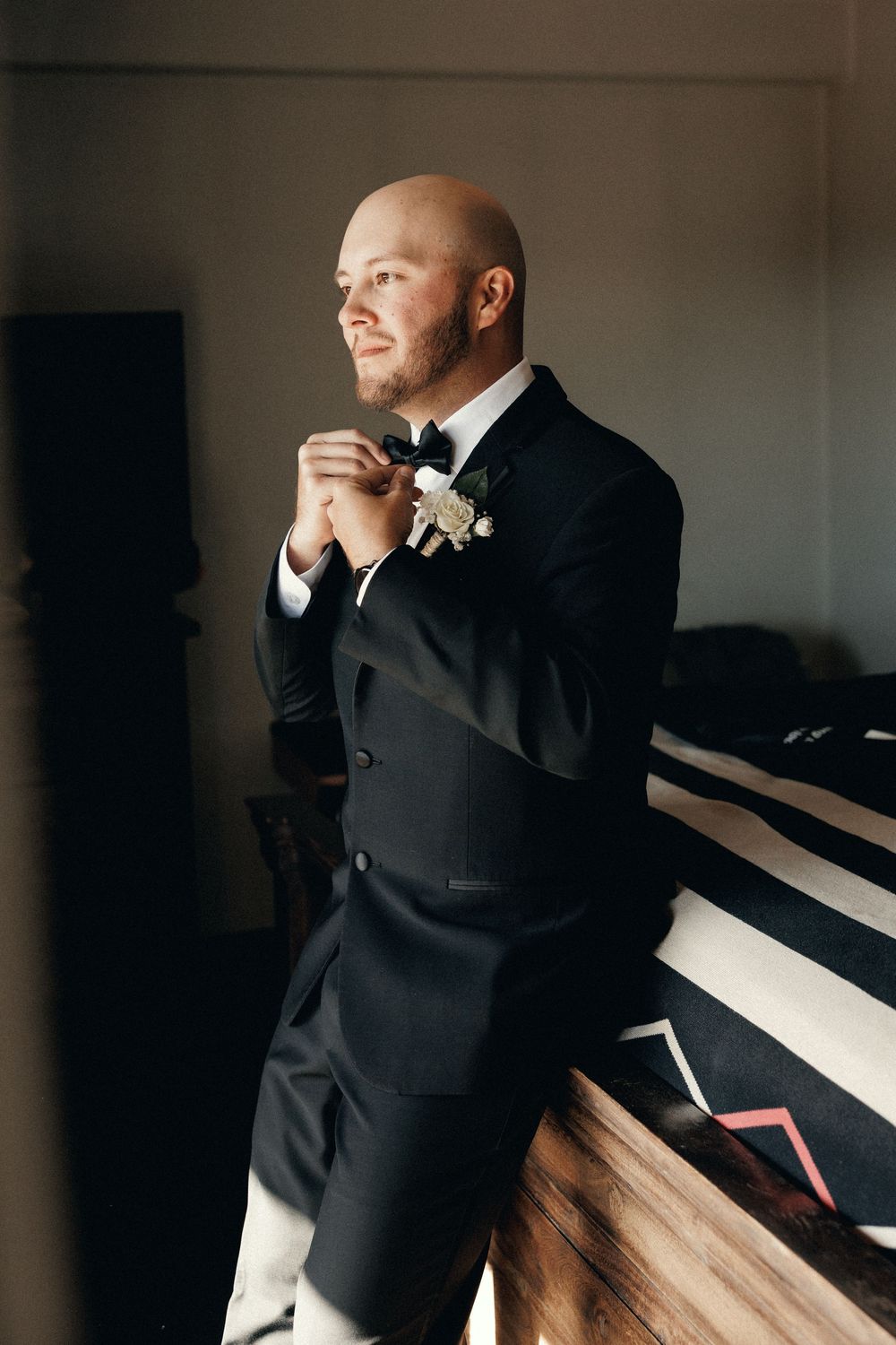 A smiling bald groom in a black tuxedo adjusting his bow tie with a white floral boutonniere.