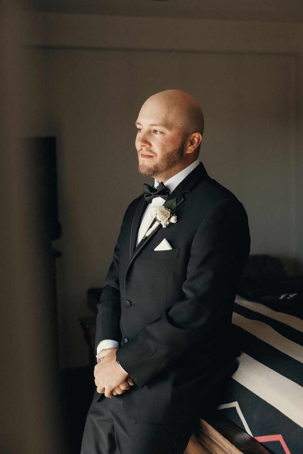 Bald groom in black tuxedo with bow tie and boutonniere posing indoors on wedding day.