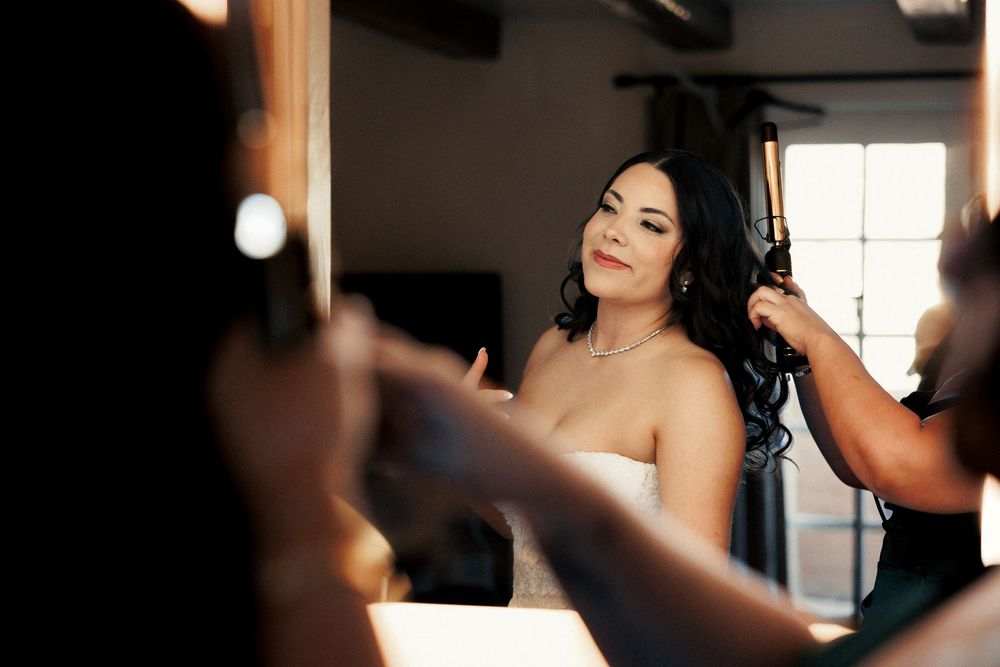 Bride getting hair styled with curling iron before wedding ceremony, wearing strapless gown and diamond necklace.