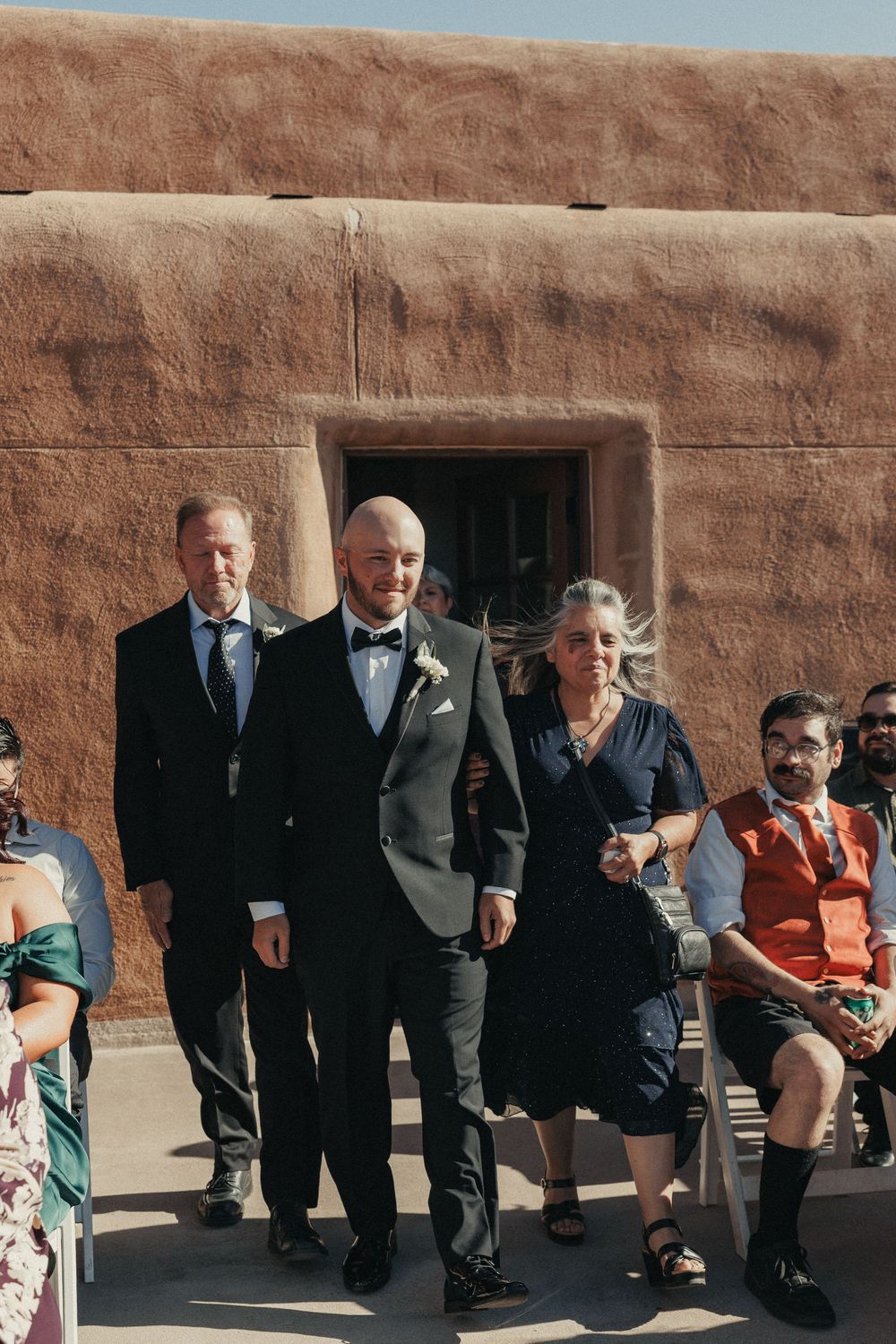 Groom escorted down outdoor aisle by parents at adobe venue wedding ceremony.