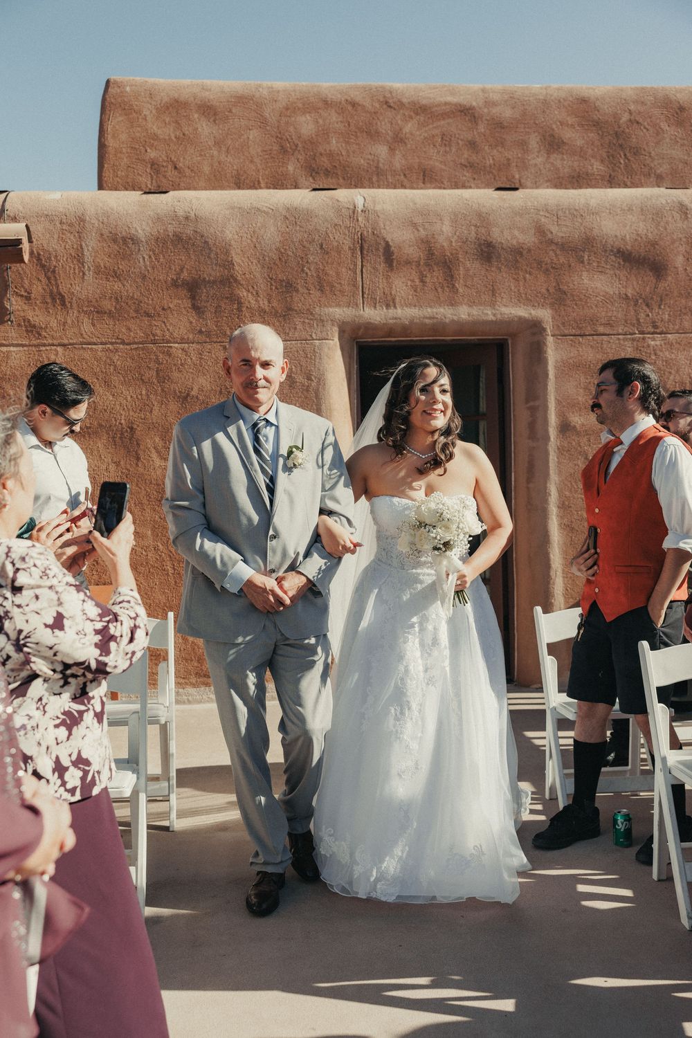 Bride in lace gown and veil walks down outdoor aisle with father at adobe venue.