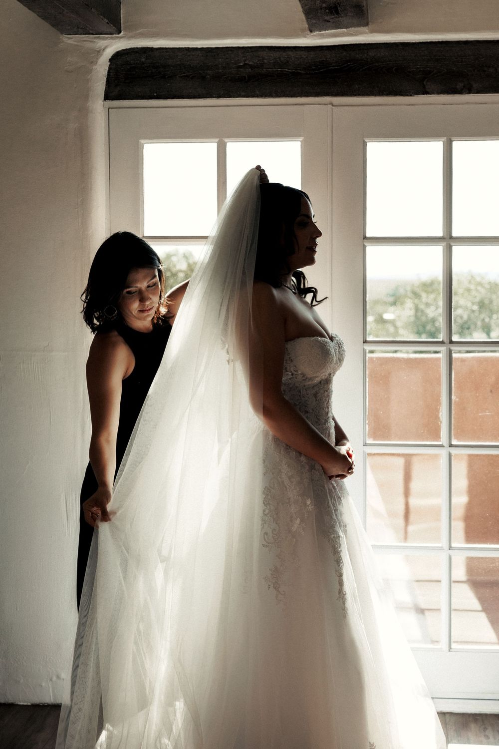 Bridesmaid helping bride with long white veil and lace wedding gown near bright windows.
