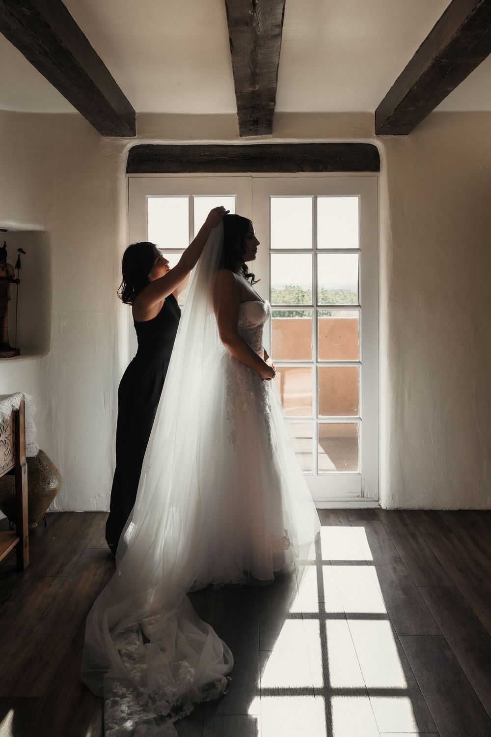 Bridesmaid in black dress helping bride with flowing white veil and gown by sunlit window in rustic room.