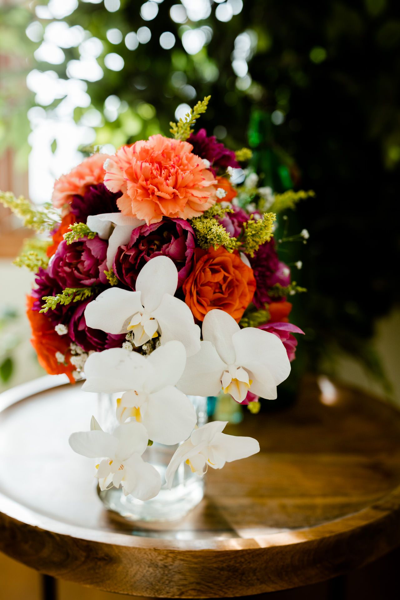 A vibrant and colorful photo of a wedding bouquet in a vase on a tabletop, White orchids and pink and purple roses with greenery.