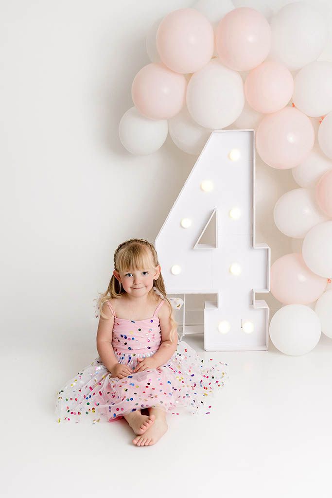 A smiling girl in a pink dress sits beside a large illuminated number 4 with pink and white balloons by Murrieta child photographer..