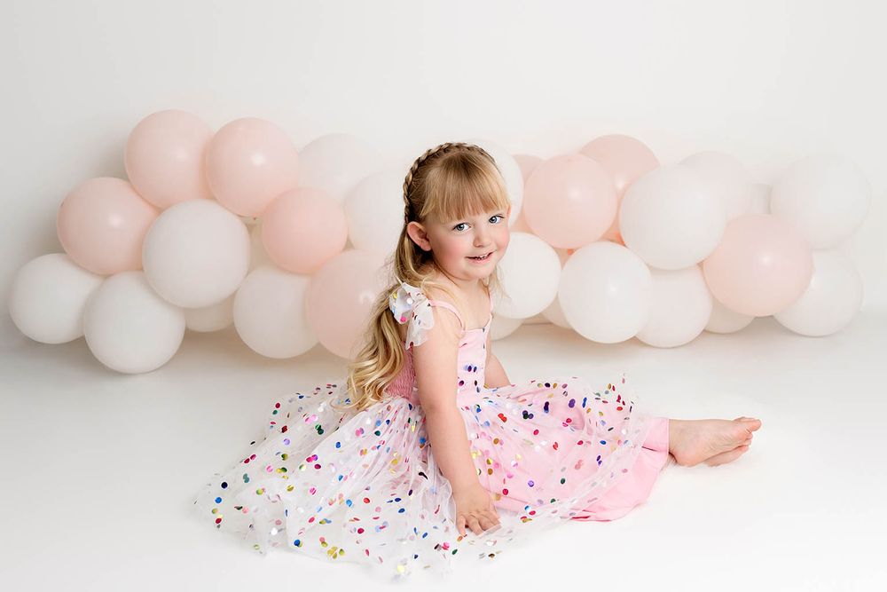 A young girl in a pink confetti dress sits gracefully in front of a pink and white balloon garland by Murrieta child photographer.