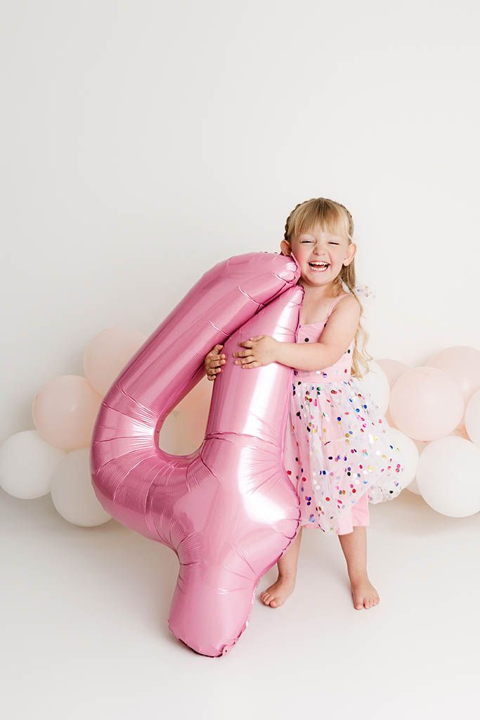 A laughing girl in a pink dress hugs a large pink number 4 balloon at her birthday photoshoot by Murrieta child photographer.