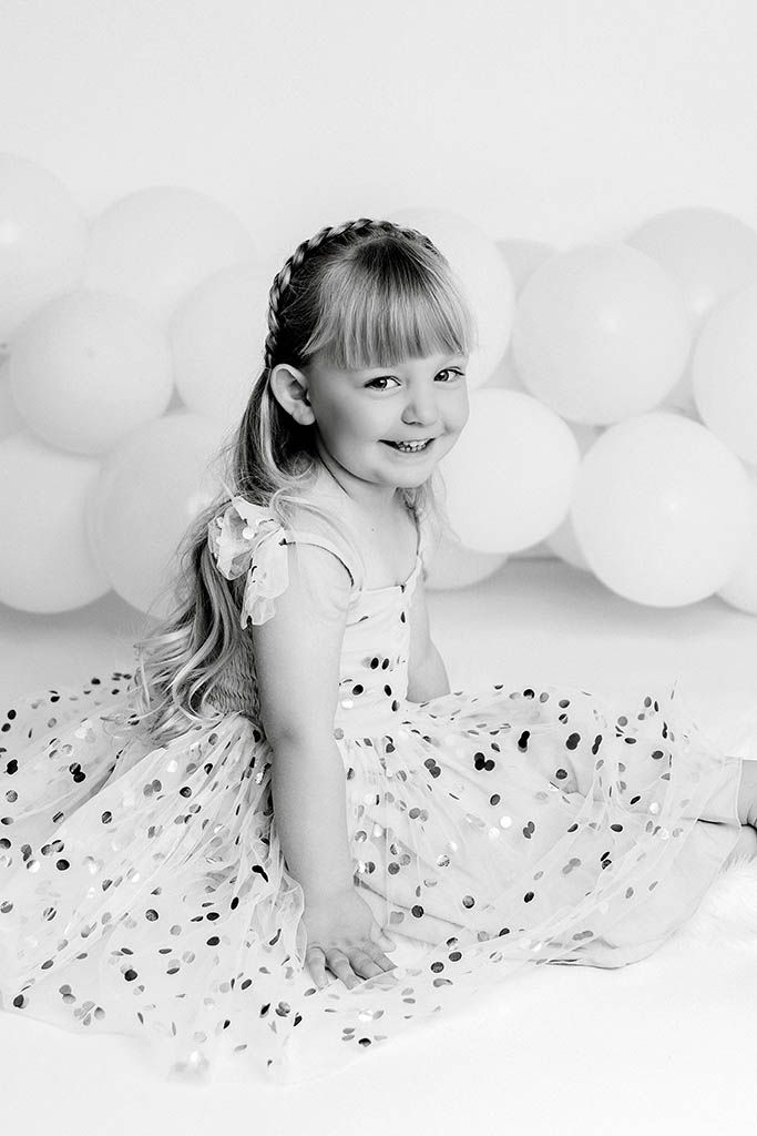 Black and white photo of a smiling girl in a dot dress sitting before balloon backdrop by Murrieta child photographer.