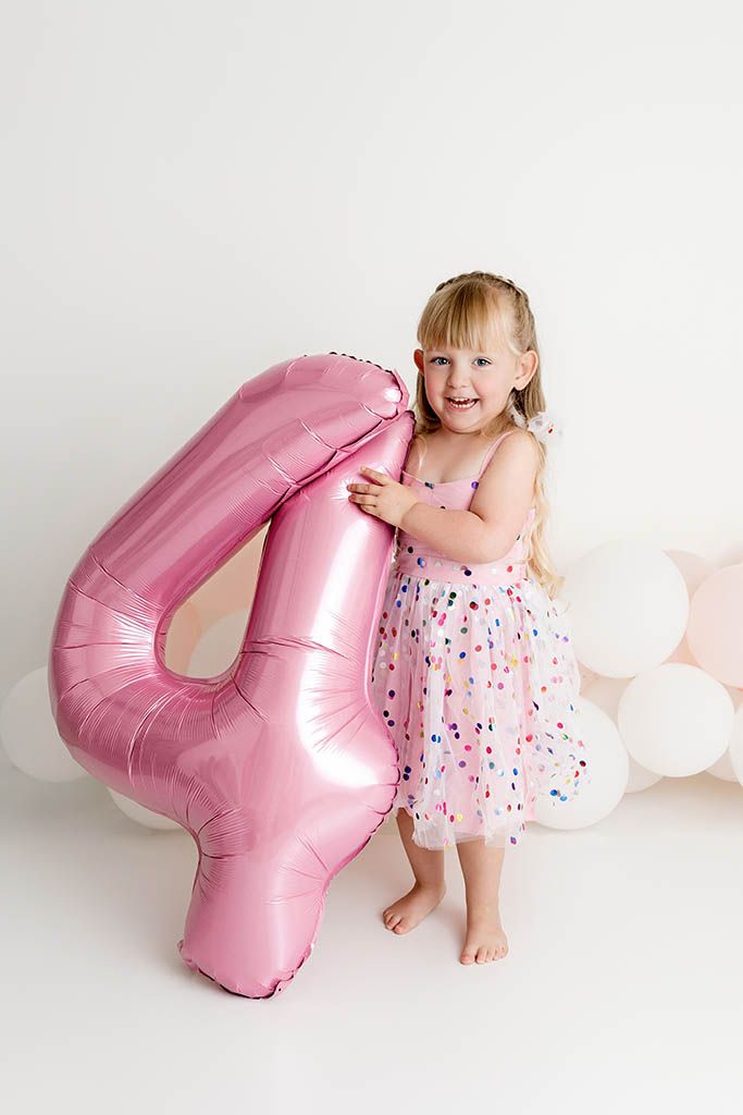 Smiling little girl hugging large pink number 4 balloon in confetti dress by Murrieta child photographer.