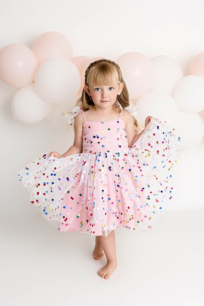 Young girl in pink confetti dress twirling barefoot in front of white and pink balloon backdrop by Murrieta child photographer.