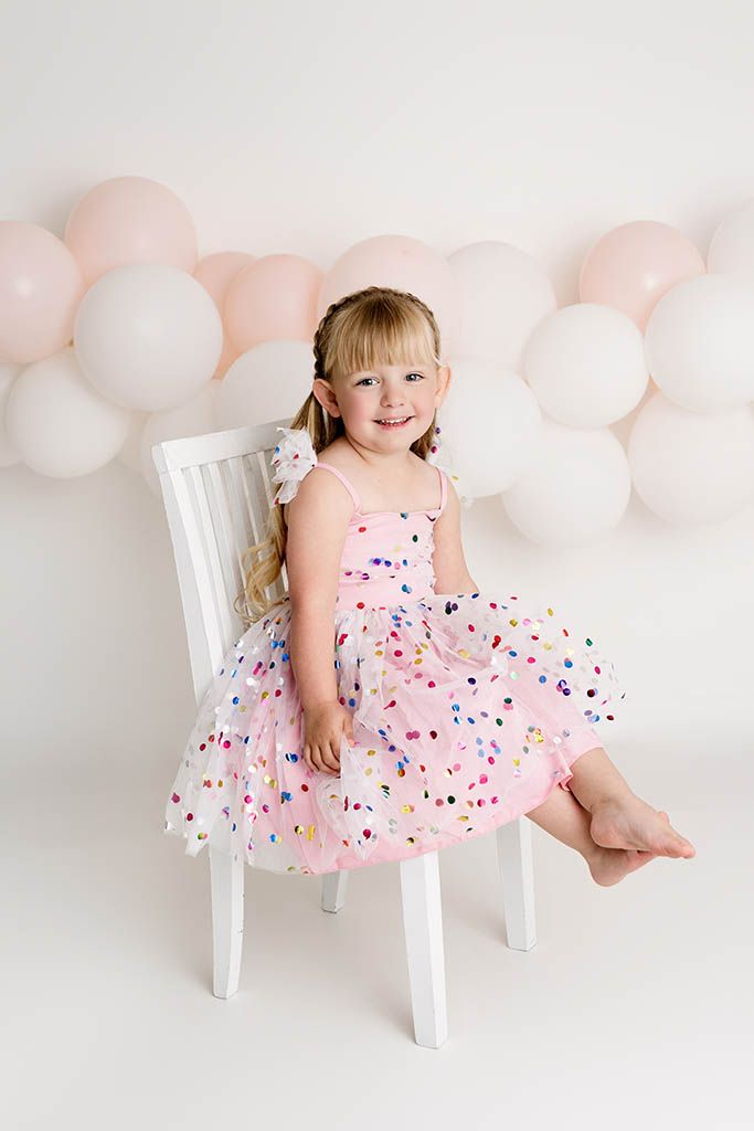 Happy young girl in pink confetti dress sitting on white chair with balloon backdrop by Murrieta child photographer.
