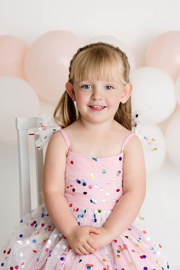 Happy young girl in a pink confetti dot dress sitting on a chair with balloon backdrop by Murrieta child photographer.