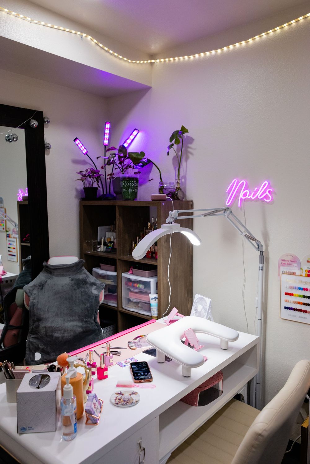 A nail salon workspace with a white desk, pink neon sign, purple LED lights, and professional manicure equipment.