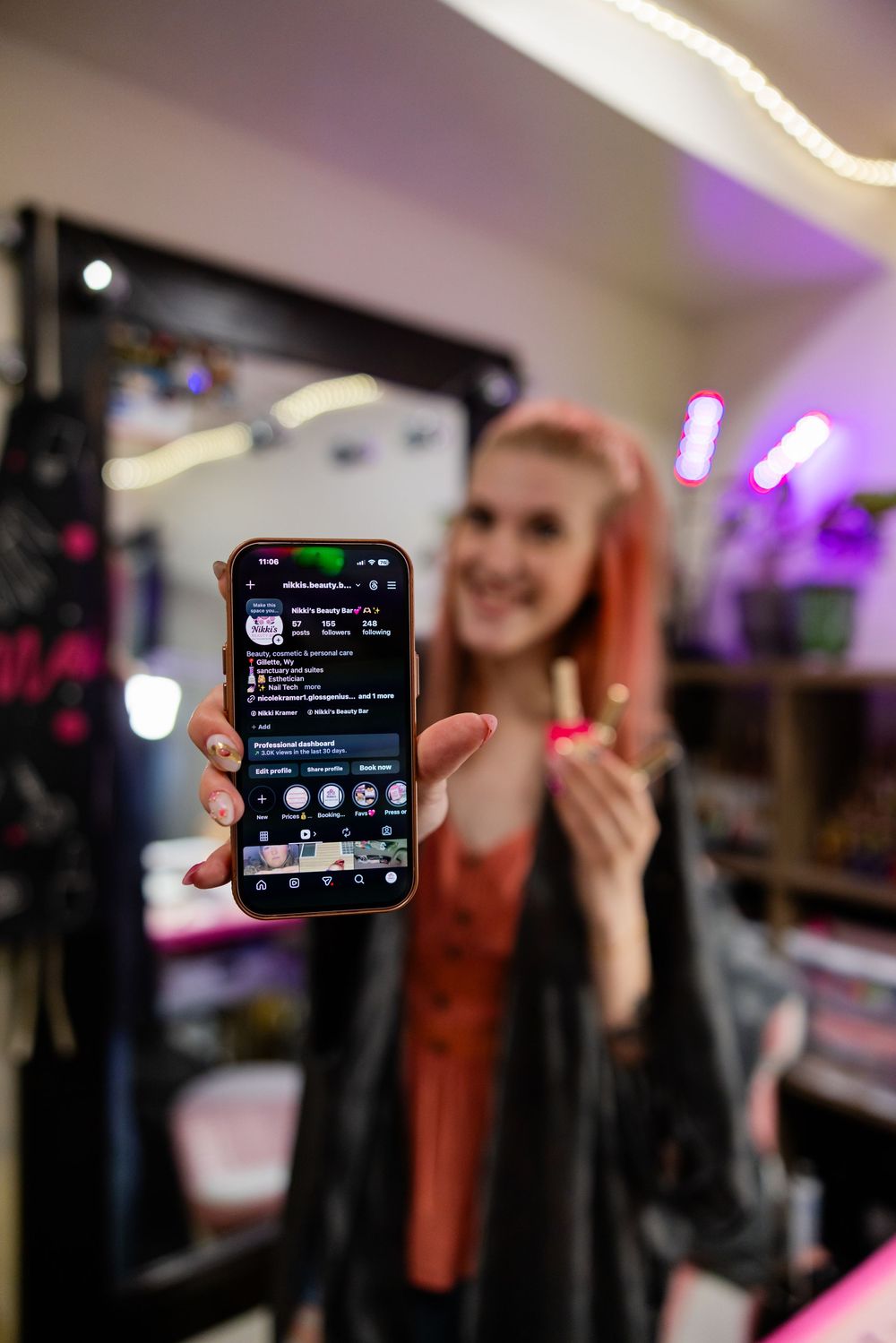 Smiling woman with pink hair holds up smartphone showing social media profile in neon-lit room.