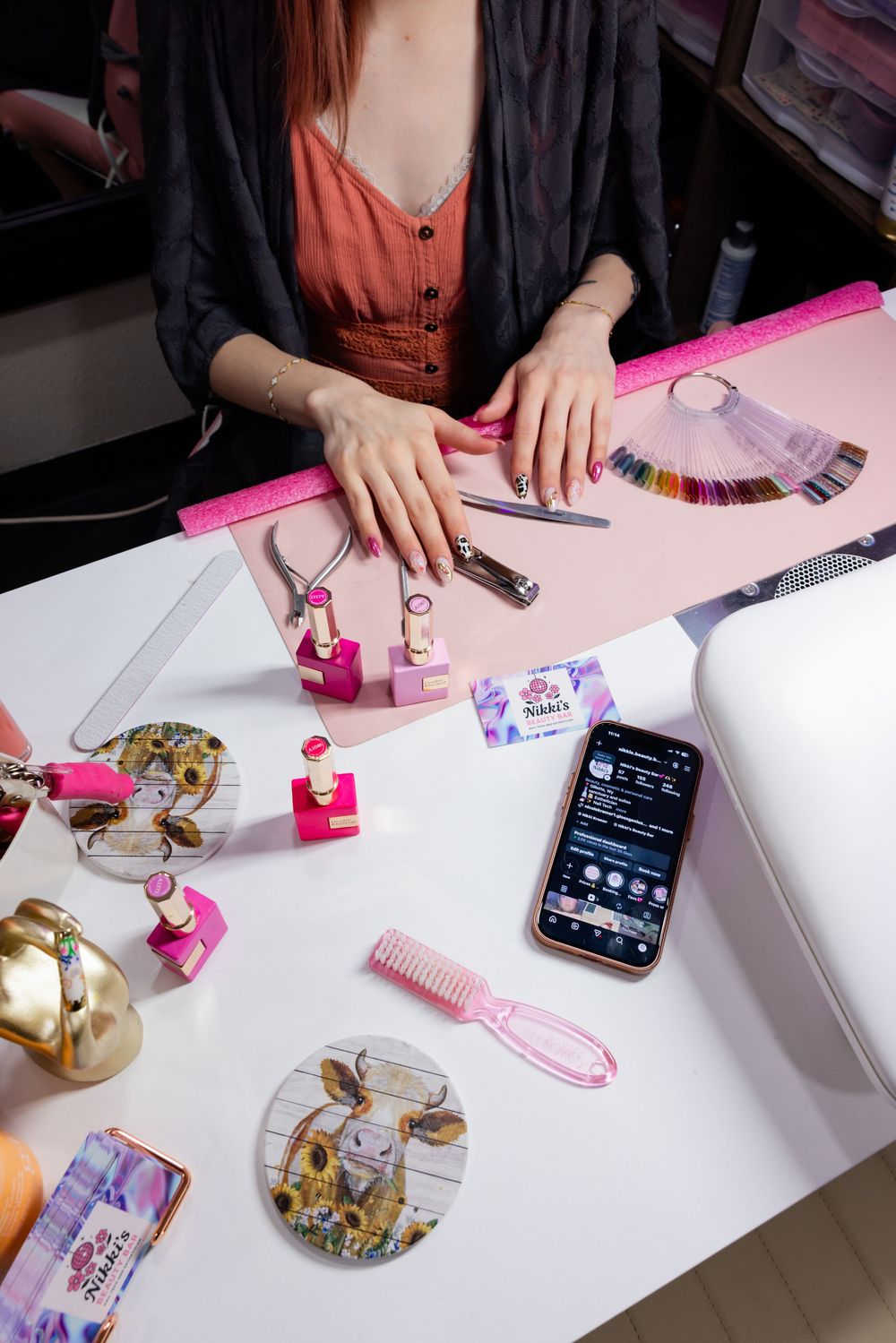 Nail technician at pink salon desk with tools, color swatches, cosmetics, and a smartphone displaying social media.