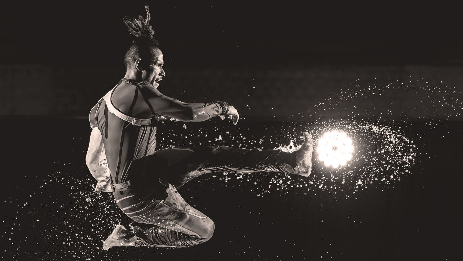 Black and white photo of a water dancer kicking with a background circular light highlighting water particles.