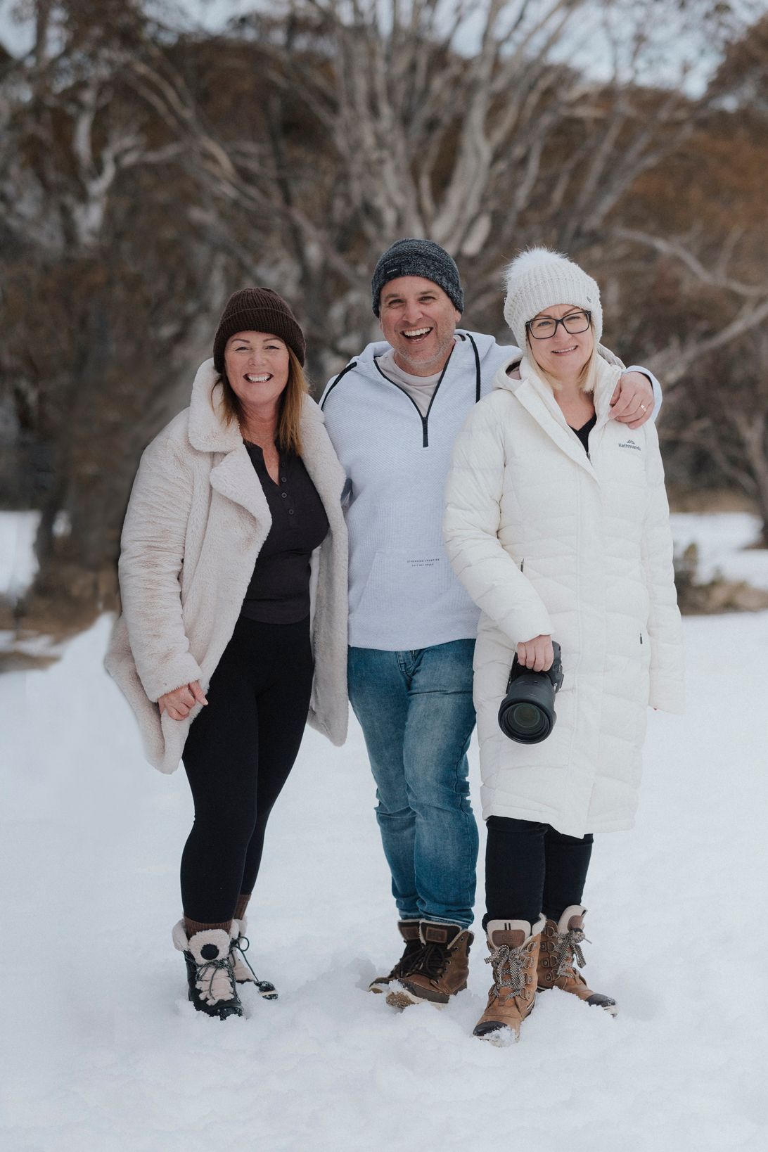 Three people smiling and posing together in a snowy winter landscape, wearing warm coats and beanies.