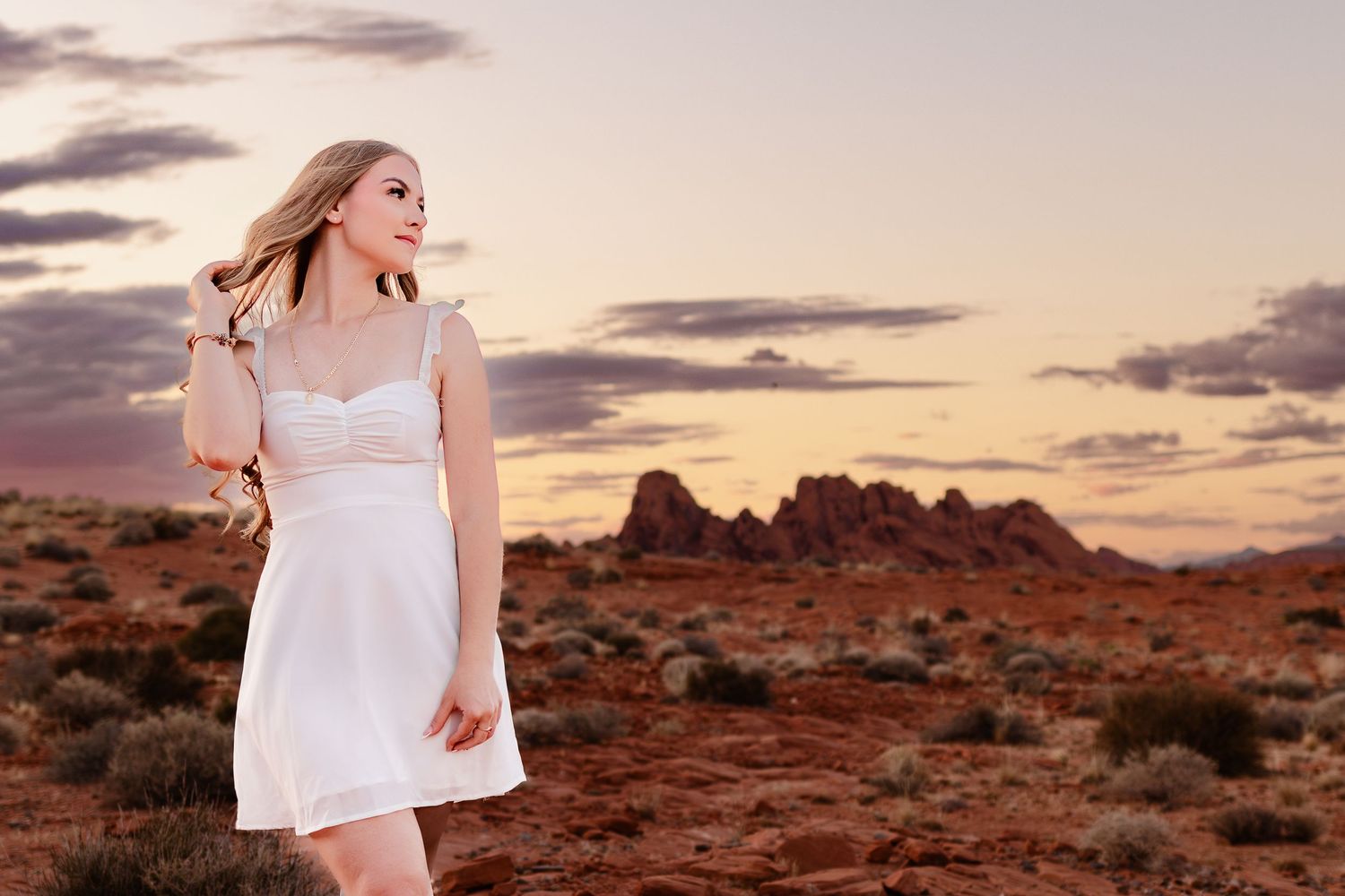 Young blonde woman in white dress poses in desert landscape at sunset with red rock formations in background.