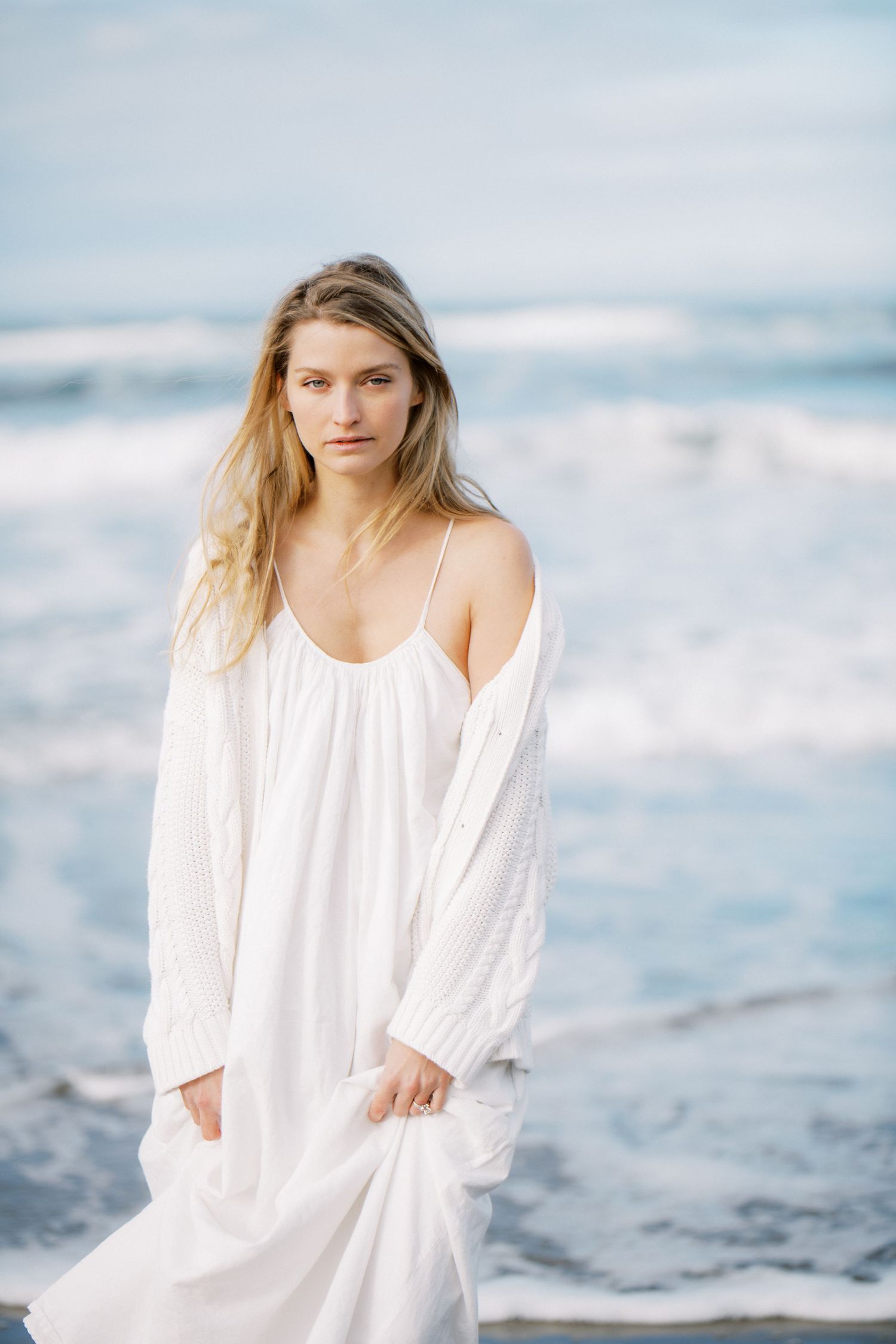 Young blonde woman in white knit cardigan poses on a beach with soft natural light and coastal rocks in background.