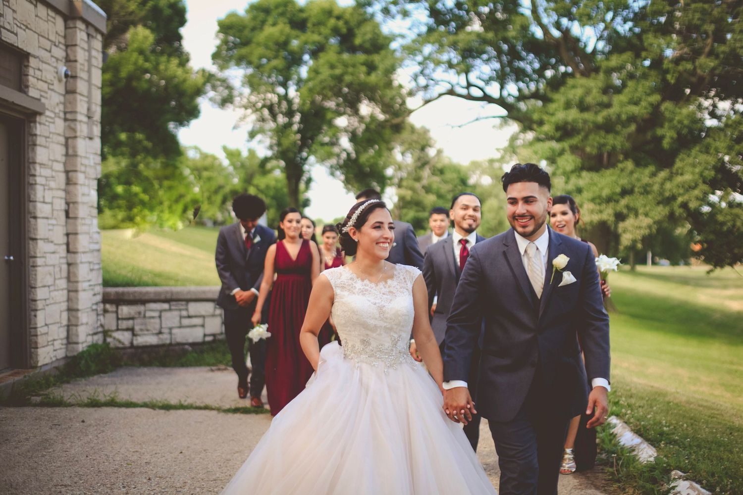 Bride in white ballgown and groom in navy suit walk hand-in-hand with wedding party outdoors near stone building.