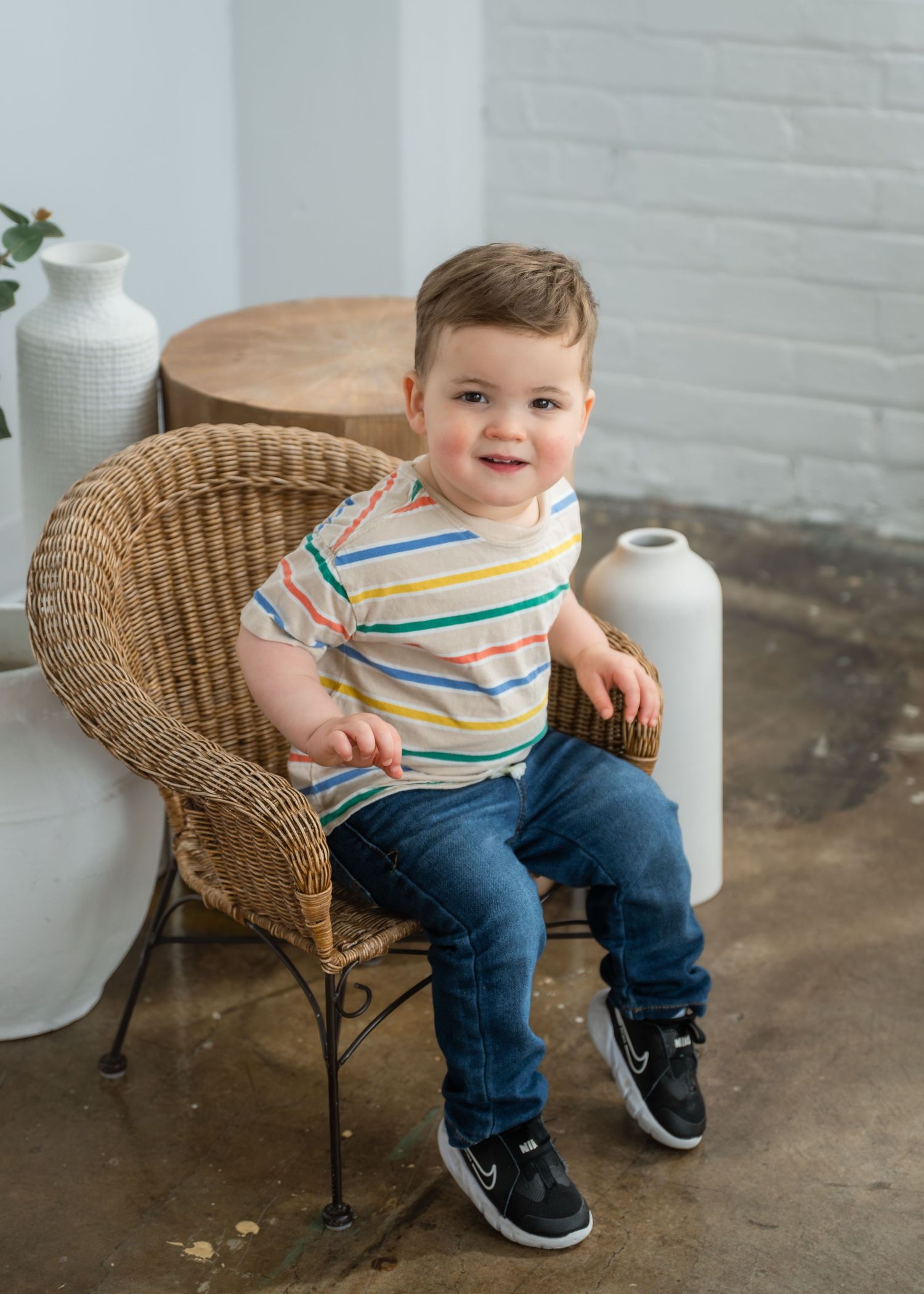 Toddler boy in striped shirt and jeans sits smiling in wicker chair with white vases in background.