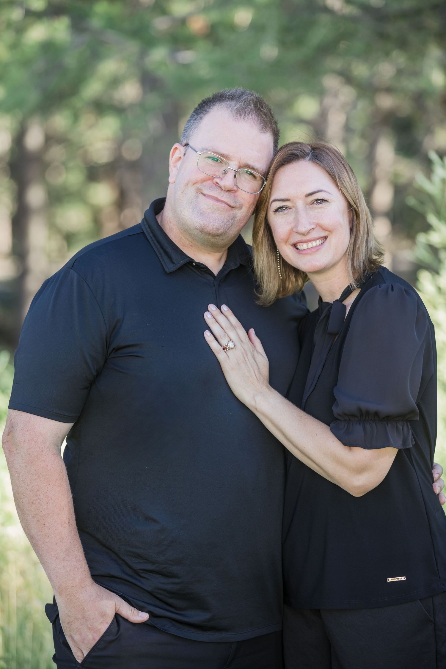 A smiling couple in matching black outfits sitting together, the man with his arm around the woman, posing for a portrait.