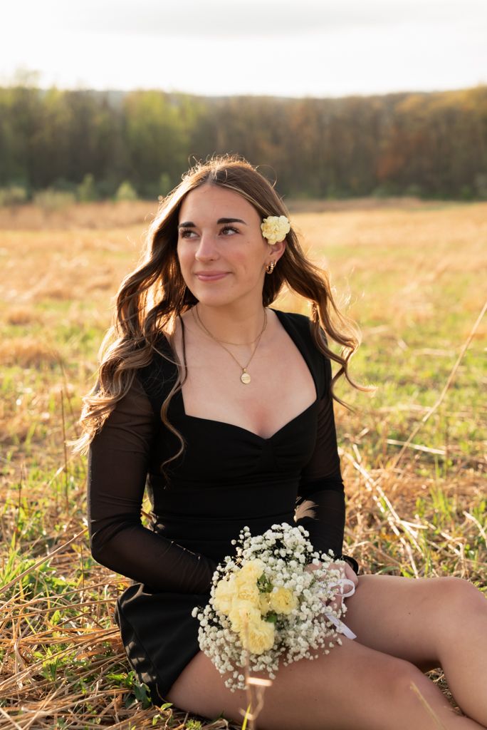 Young woman in black dress holding white flowers sits in golden field with flower in her hair.