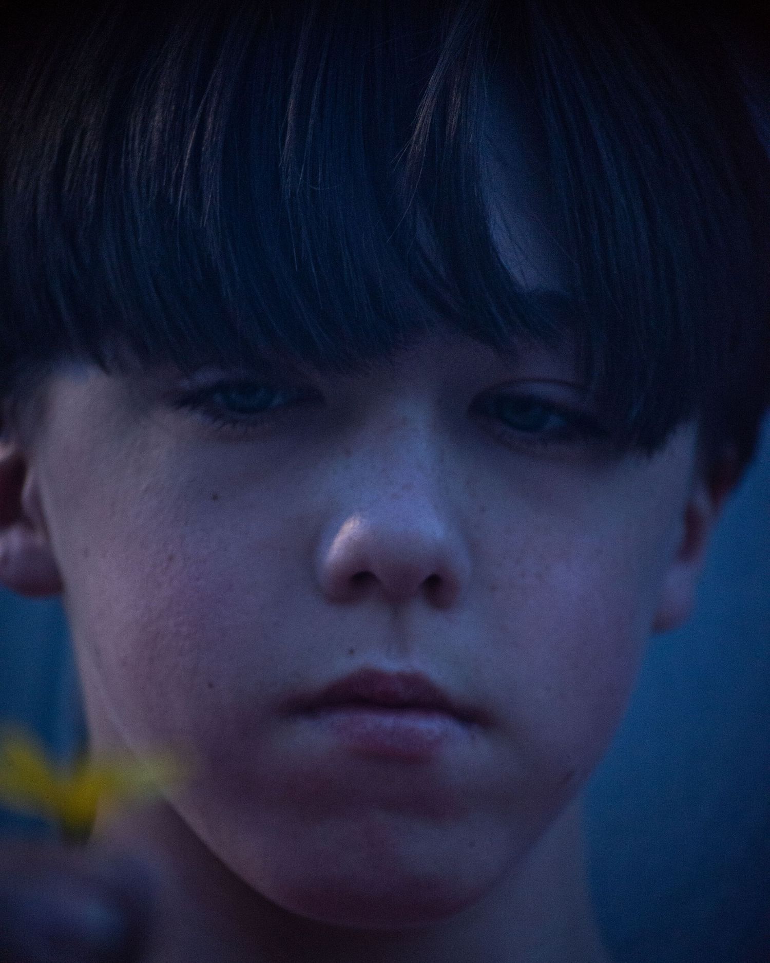 Close-up portrait of a young boy with dark hair, freckles, and blue eyes in dramatic moody lighting.
