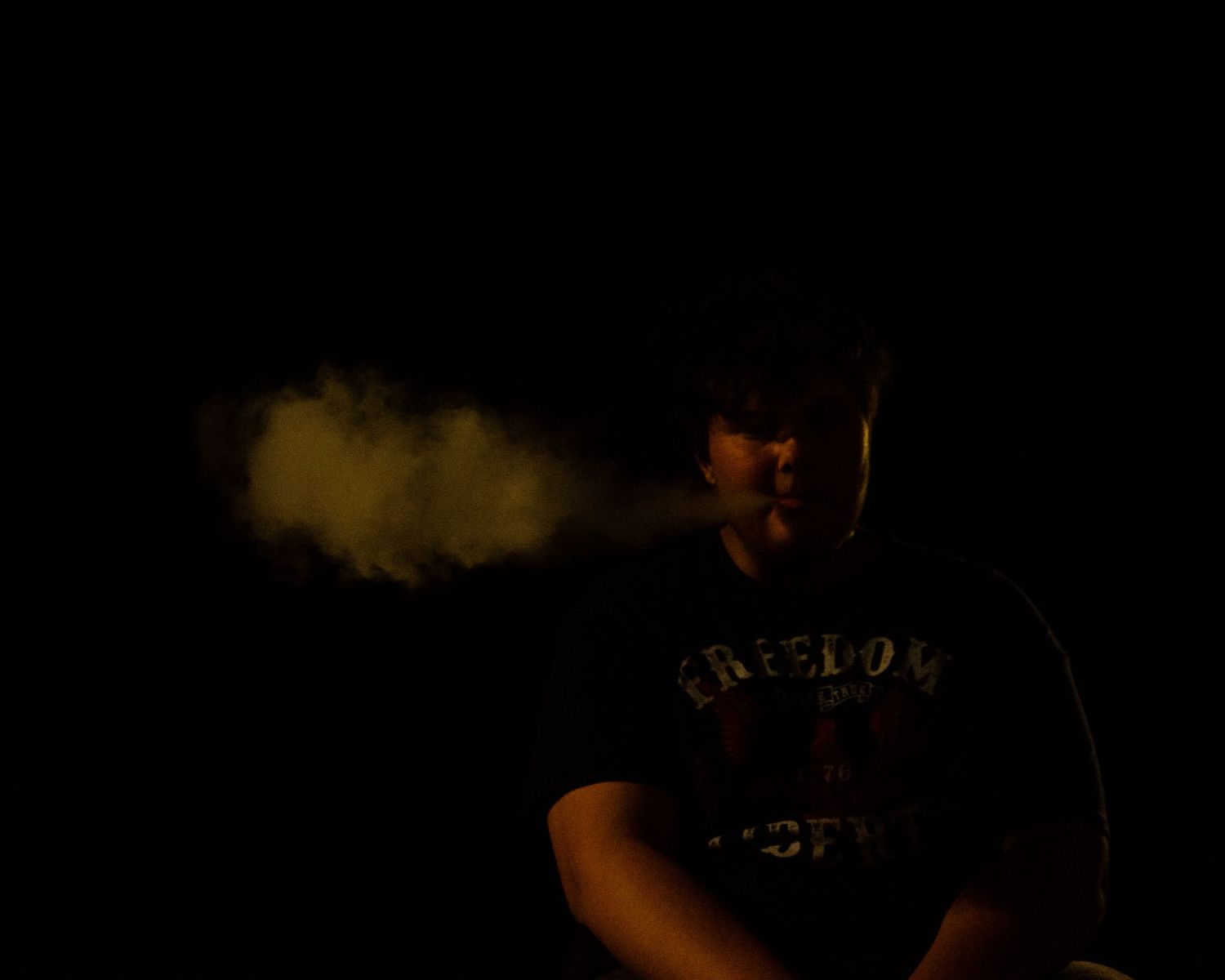 Man in dark setting exhaling vapor cloud from e-cigarette, wearing a black graphic tee shirt.