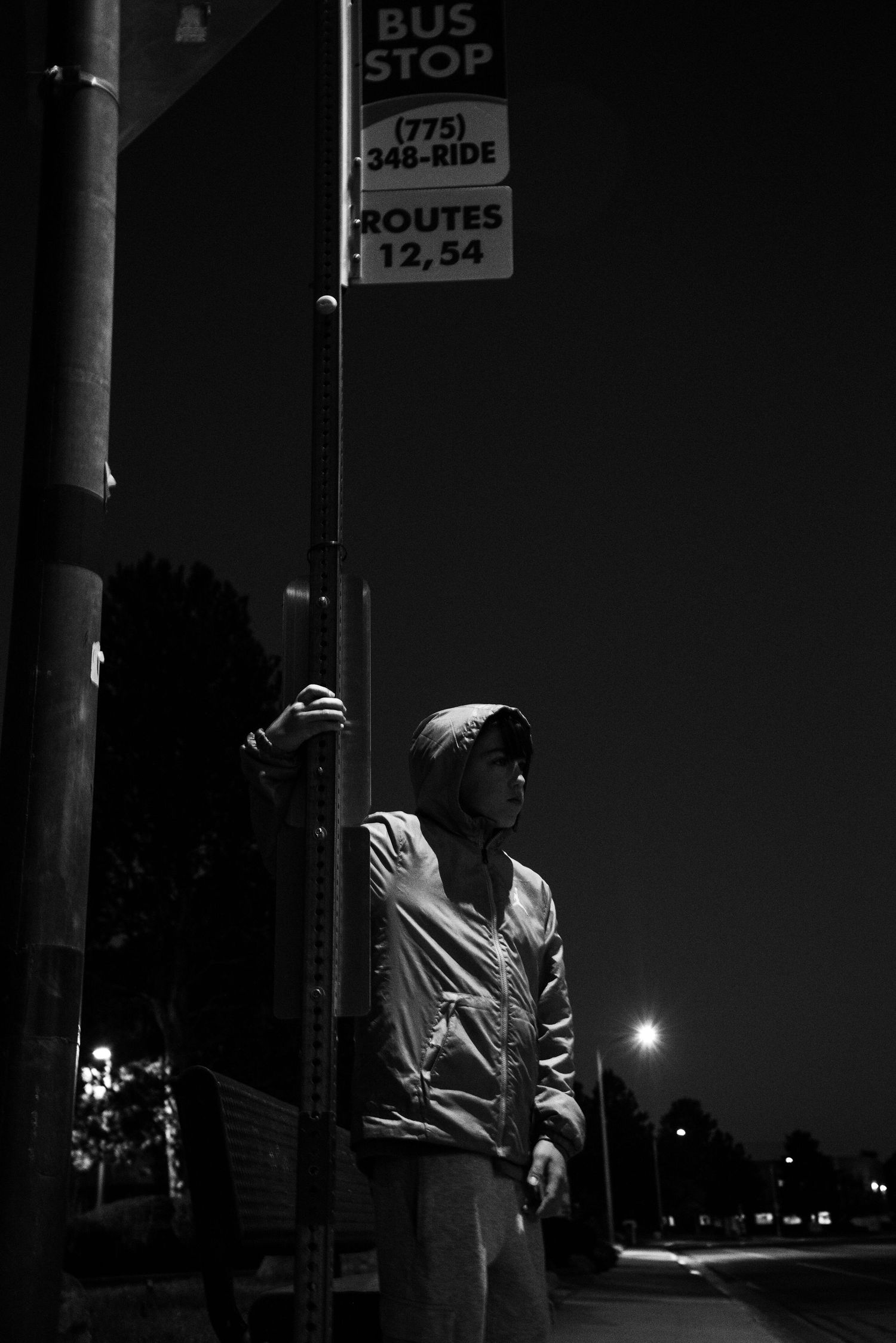 Person in hooded jacket standing at a bus stop at night, routes 12 and 54, black and white photo.