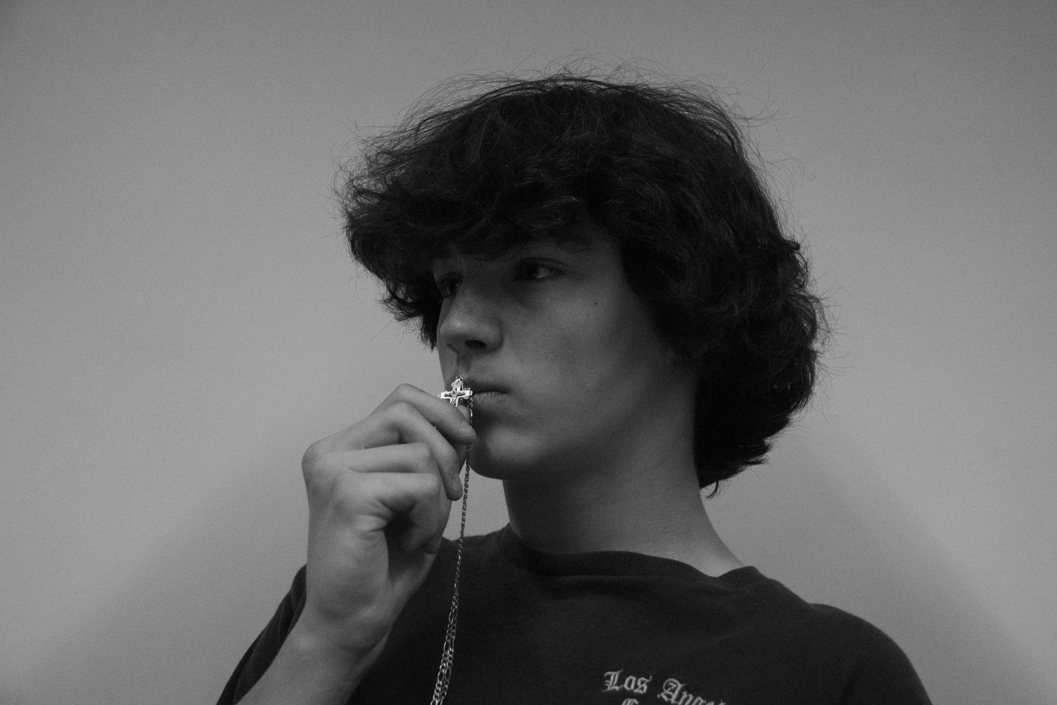 Black and white portrait of a young man with curly hair holding a necklace to his lips, wearing a graphic tee.