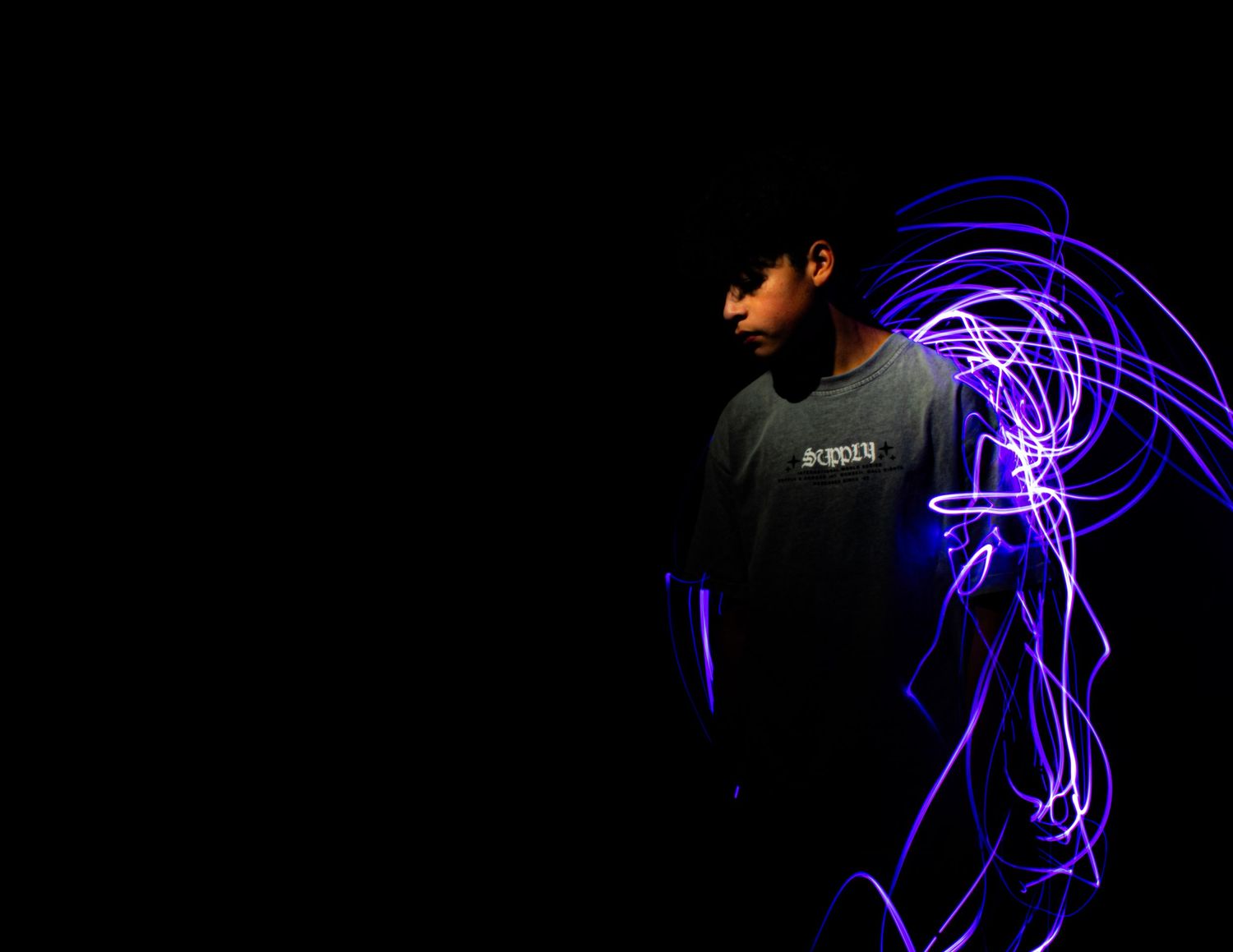 Person standing against dark background surrounded by vibrant blue and purple light painting trails in long exposure photography.