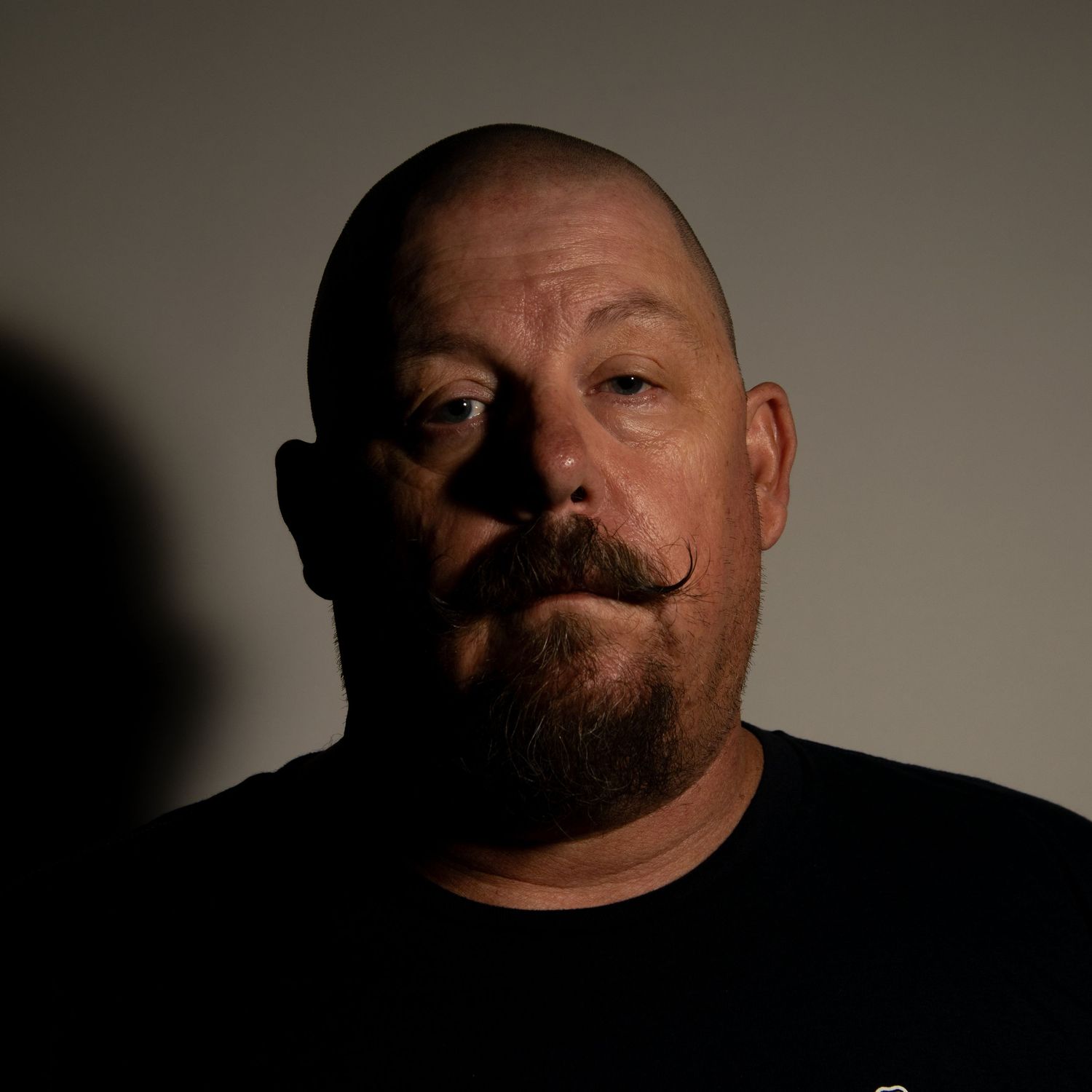 Bald man with a styled mustache and goatee wearing a black shirt, photographed in dramatic low-key lighting.