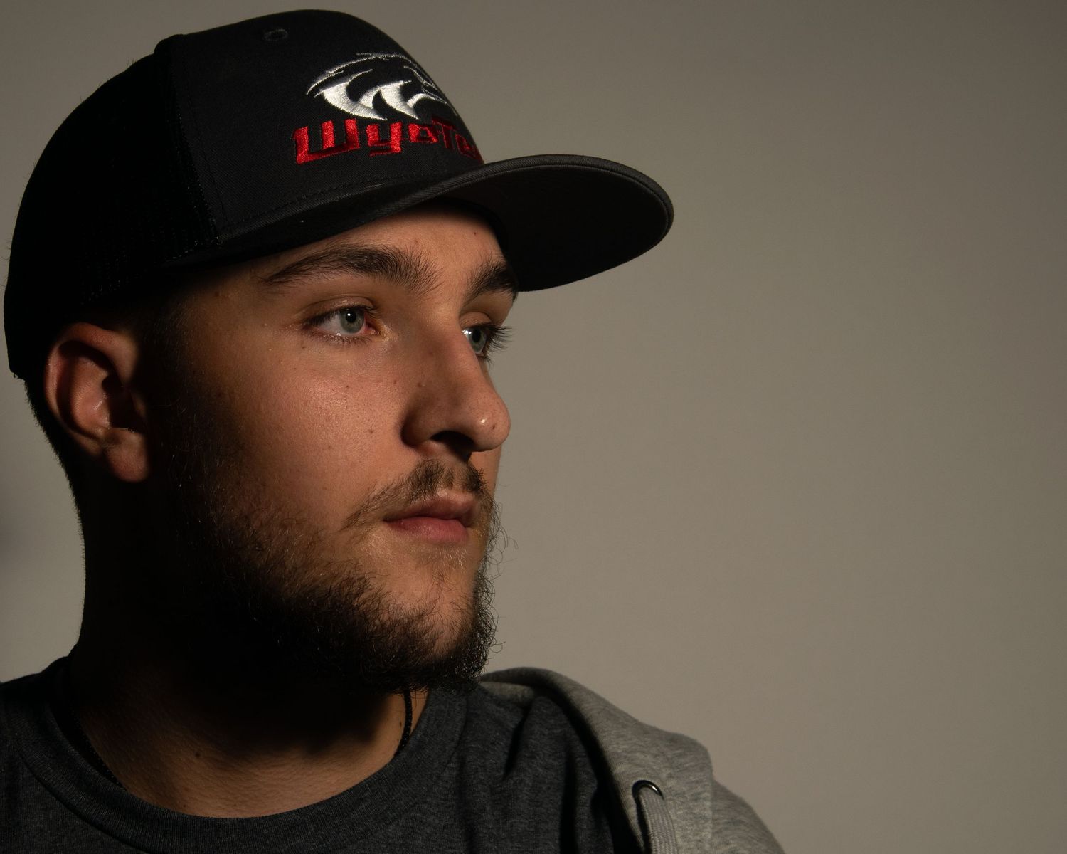 Young man with beard wearing a black snapback cap looks thoughtfully to the side in a studio portrait.